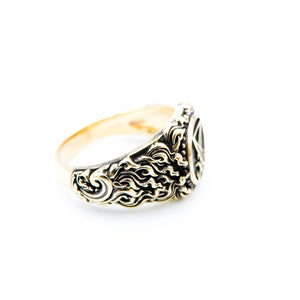 May include: A gold-coloured ring with detailed, swirling patterns and a central design. The ring's band is smooth, while the face features ornate carvings. The ring is set against a clean white background, highlighting its design.