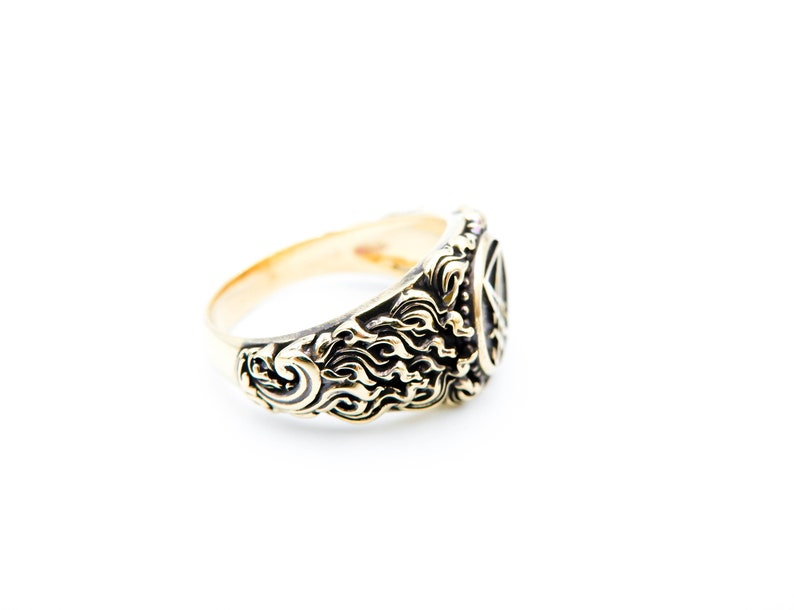 May include: A gold-coloured ring with detailed, swirling patterns and a central design. The ring's band is smooth, while the face features ornate carvings. The ring is set against a clean white background, highlighting its design.