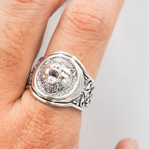 K&ouml;nnte beinhalten: Ein silberner Ring mit einem detaillierten B&auml;renkopfdesign auf der Vorderseite und einem detaillierten Blumendesign auf der Band.