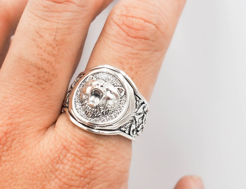 K&ouml;nnte beinhalten: Ein silberner Ring mit einem detaillierten B&auml;renkopfdesign auf der Vorderseite und einem detaillierten Blumendesign auf der Band.