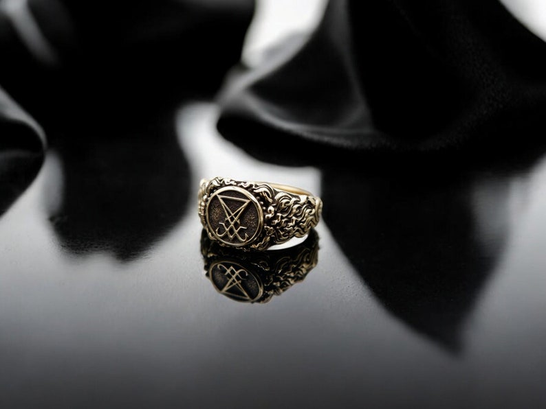 May include: A gold ring with an intricate design. The ring has a central emblem within a circular frame, surrounded by ornate patterns. The ring is set against a dark, reflective surface, creating a dramatic contrast.
