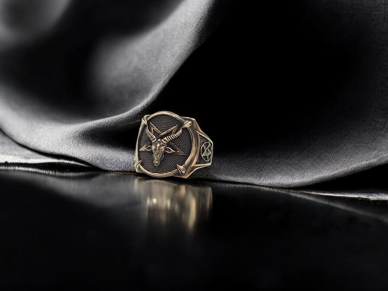 May include: A gold ring with a black enamel design featuring a goat's head with horns and a pentagram.