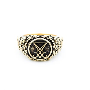 May include: A gold-coloured ring with an ornate floral design around the band. The ring features a central emblem within a circle, with a dark background and a stylized symbol. The ring is set against a white background.