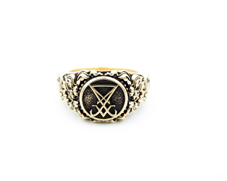 May include: A gold-coloured ring with an ornate floral design around the band. The ring features a central emblem within a circle, with a dark background and a stylized symbol. The ring is set against a white background.