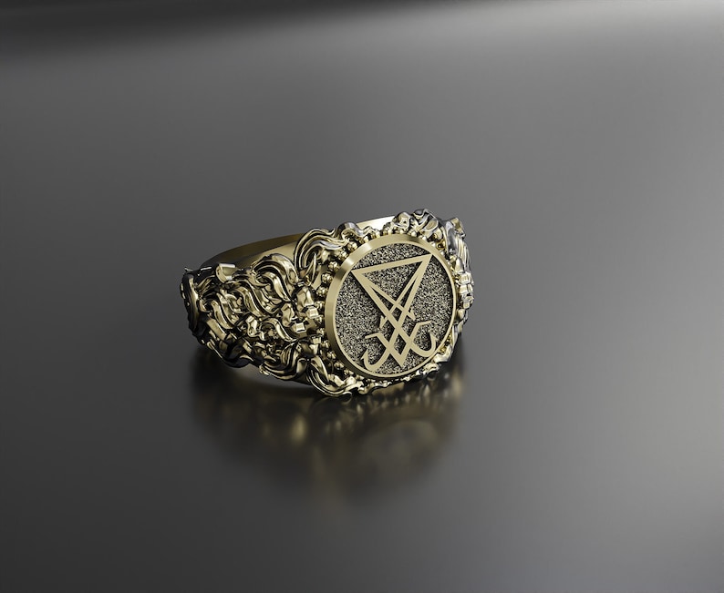 May include: Gold-coloured ring with an elaborate, ornate design. The ring has a central emblem within a circular frame, surrounded by intricate floral and scrollwork. The emblem is a stylized symbol with a triangle and lines.