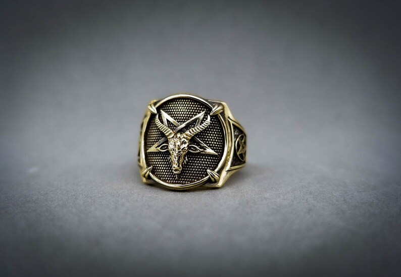 May include: A gold-toned ring with a pentagram design featuring a goat's head in the center. The ring has a textured surface and a detailed design.
