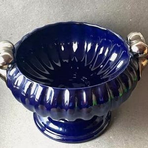 Peut inclure: Un bol en céramique bleu foncé avec un extérieur cannelé et des poignées argentées. Le bol a une large ouverture et repose sur une petite base ronde. L'intérieur reflète la lumière.