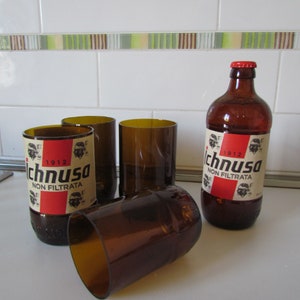 Puede incluir: Una botella de cerveza de vidrio marrón y cuatro vasos a juego. La botella tiene una etiqueta roja y blanca con el texto "Ichnusa Non Filtrata". Los vasos están hechos de botellas de cerveza cortadas.