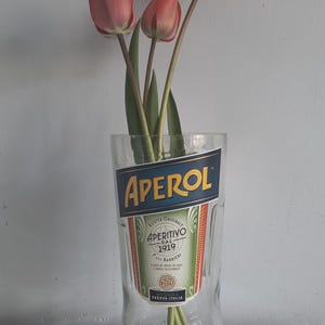 May include: A glass with the Aperol logo, holding several pink tulips. The glass has the words "APEROL" and "APERITIVO DAL 1919" printed on it. The tulips have green stems and leaves.