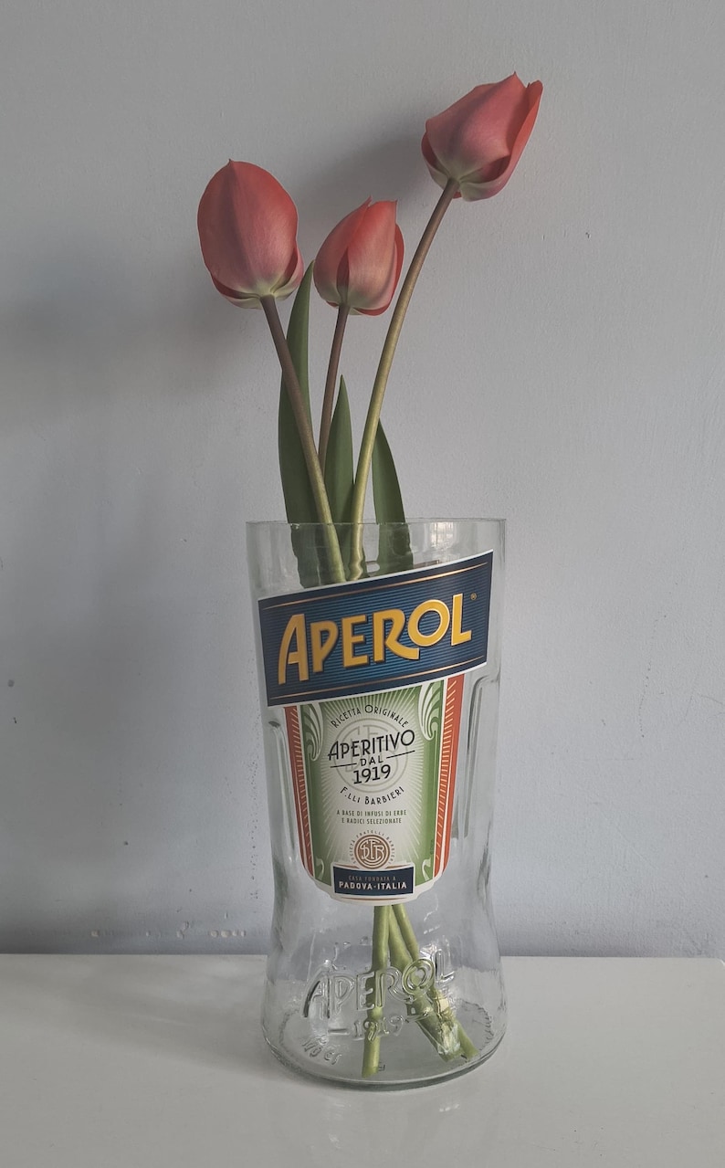 May include: A glass with the Aperol logo, holding several pink tulips. The glass has the words "APEROL" and "APERITIVO DAL 1919" printed on it. The tulips have green stems and leaves.