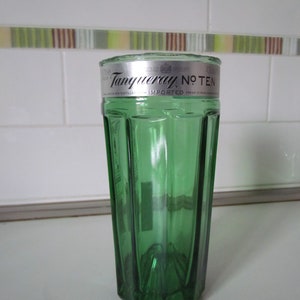 May include: A green glass tumbler with a silver rim. The tumbler has a faceted design and the words "Tanqueray No. TEN" are printed on the silver rim. The glass is sitting on a white surface.