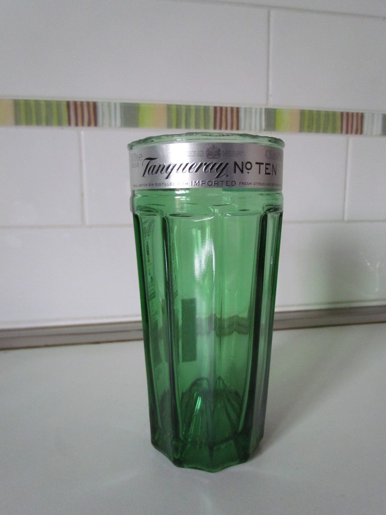 May include: A green glass tumbler with a silver rim. The tumbler has a faceted design and the words "Tanqueray No. TEN" are printed on the silver rim. The glass is sitting on a white surface.