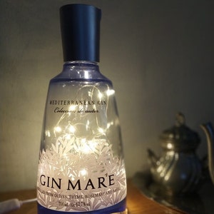 May include: A clear glass bottle with a blue label that says "Gin Mare" and "Mediterranean Gin". The bottle is filled with white fairy lights and is sitting on a wooden surface.
