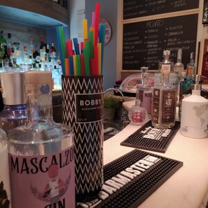 May include: A bar scene with bottles of liquor, including Mascalzo Gin and Bobby's Schiedam Dry Gin. A container of colorful straws sits next to the bottles. Black bar mats and a chalkboard menu are also visible.