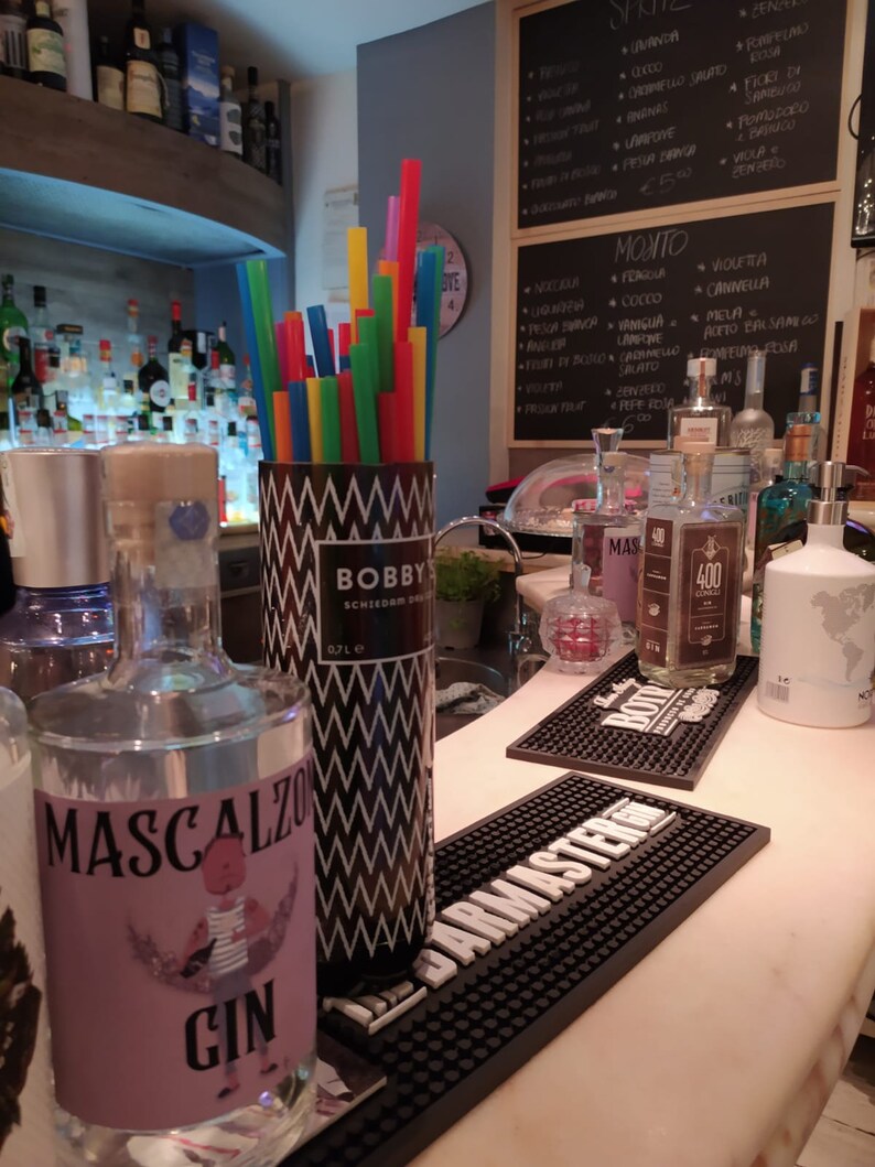 May include: A bar scene with bottles of liquor, including Mascalzo Gin and Bobby's Schiedam Dry Gin. A container of colorful straws sits next to the bottles. Black bar mats and a chalkboard menu are also visible.