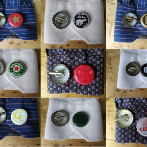 Può includere: Nove diverse paia di gemelli realizzati con tappi di bottiglia. Ogni paio presenta un design diverso, tra cui una stella rossa, un orso, il numero 15, un logo Heineken, un cerchio rosso, un quadrato verde, una lettera C gialla, un design verde e una corona con la parola "CERES".