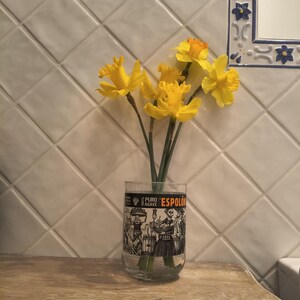 May include: A clear glass with a black and white illustration of people and the text "ESPOLON" holds a bouquet of yellow daffodils. The vase sits on a wooden surface against a white tiled wall.