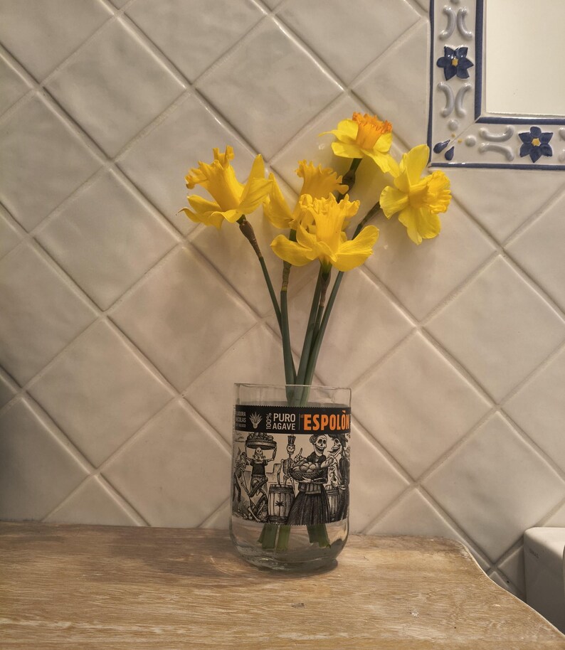 May include: A clear glass with a black and white illustration of people and the text "ESPOLON" holds a bouquet of yellow daffodils. The vase sits on a wooden surface against a white tiled wall.