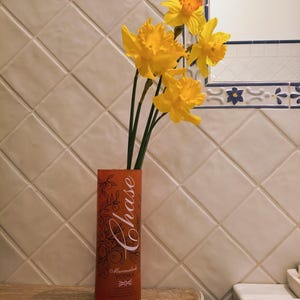 May include: An orange Chase Marmalade Vodka bottle with a floral design holds several yellow daffodils. The bottle is on a wooden surface, with a tiled wall and a mirror in the background.