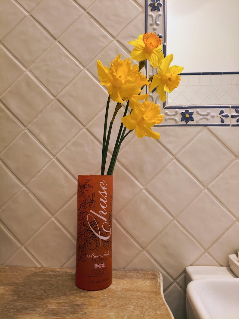 May include: An orange Chase Marmalade Vodka bottle with a floral design holds several yellow daffodils. The bottle is on a wooden surface, with a tiled wall and a mirror in the background.