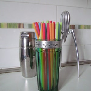 May include: A green glass with colorful straws, a stainless steel cocktail shaker, and a silver citrus juicer. The glass has the text "Tanqueray No. TEN" on it. The straws are various colors.