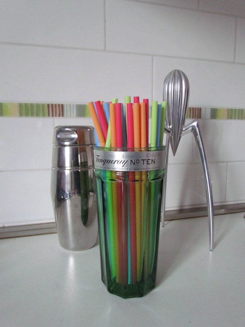 May include: A green glass with colorful straws, a stainless steel cocktail shaker, and a silver citrus juicer. The glass has the text "Tanqueray No. TEN" on it. The straws are various colors.