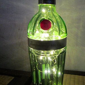 May include: A green glass bottle with the words "Tanqueray No. TEN" on a black label, illuminated by small white lights inside. The bottle has a black cap and a red circular emblem. The bottle is on a wooden surface.