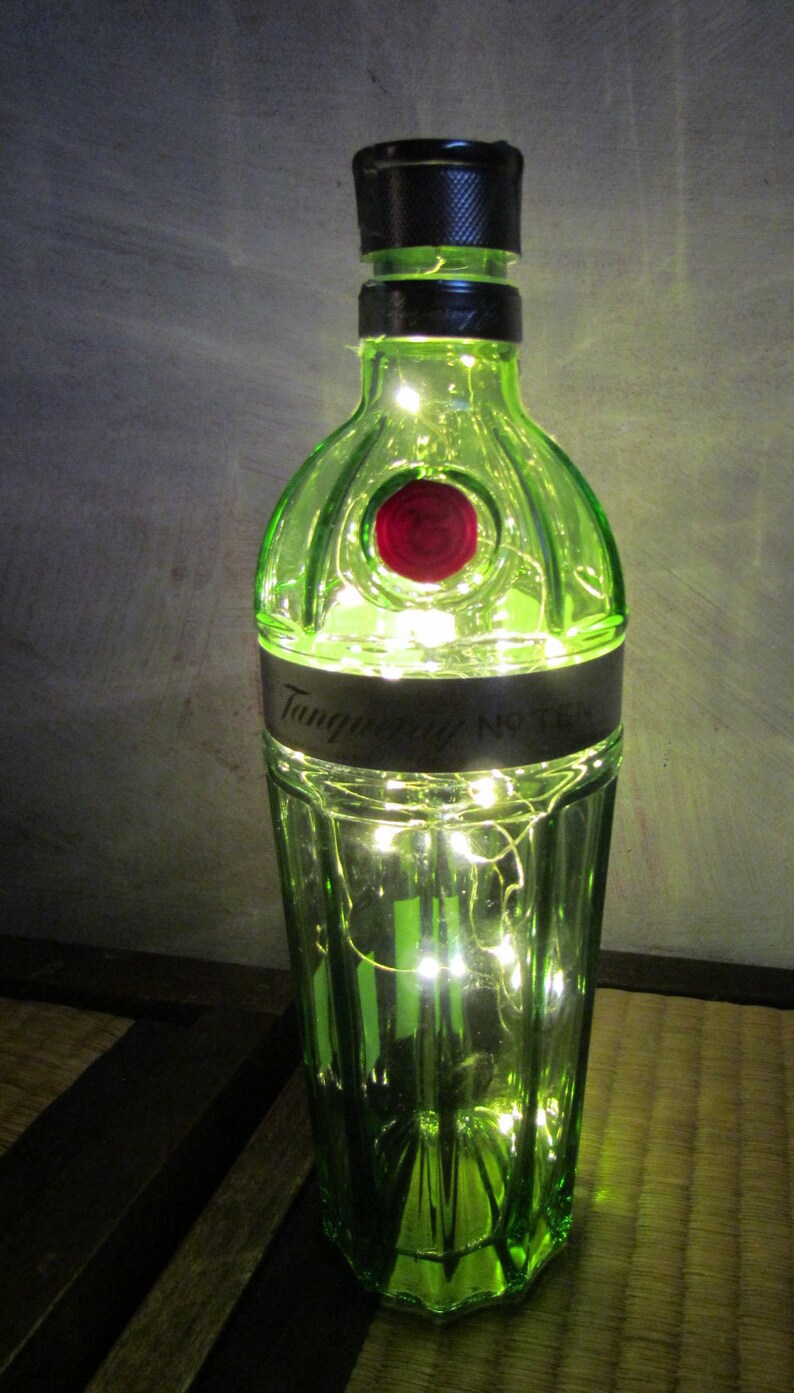 May include: A green glass bottle with the words "Tanqueray No. TEN" on a black label, illuminated by small white lights inside. The bottle has a black cap and a red circular emblem. The bottle is on a wooden surface.