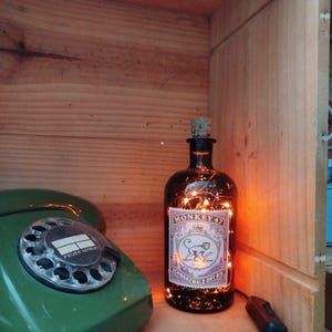 May include: A dark glass bottle with a cork stopper and a label that reads "Monkey 47 Schwarzwald Dry Gin." The bottle is illuminated with small fairy lights. A vintage green rotary dial telephone is to the left.