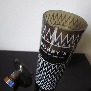 May include: A clear glass bottle with a black and white zig-zag pattern and the text "BOBBY'S Schiedam Dry Gin". The bottle is partially open, with the stopper lying next to it. The gin is 42% alcohol by volume.
