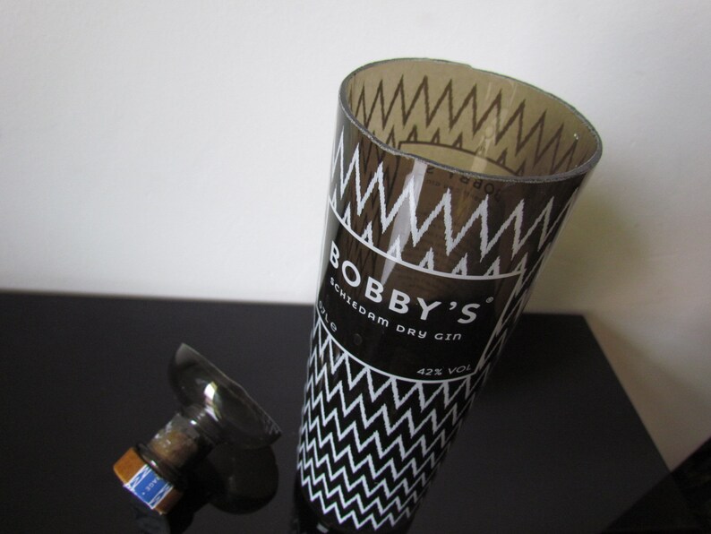 May include: A clear glass bottle with a black and white zig-zag pattern and the text "BOBBY'S Schiedam Dry Gin". The bottle is partially open, with the stopper lying next to it. The gin is 42% alcohol by volume.