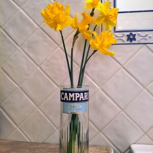 May include: A clear glass vase with a Campari label holds several bright yellow daffodils. The vase is on a wooden surface, and the background features white tiles and a mirror.