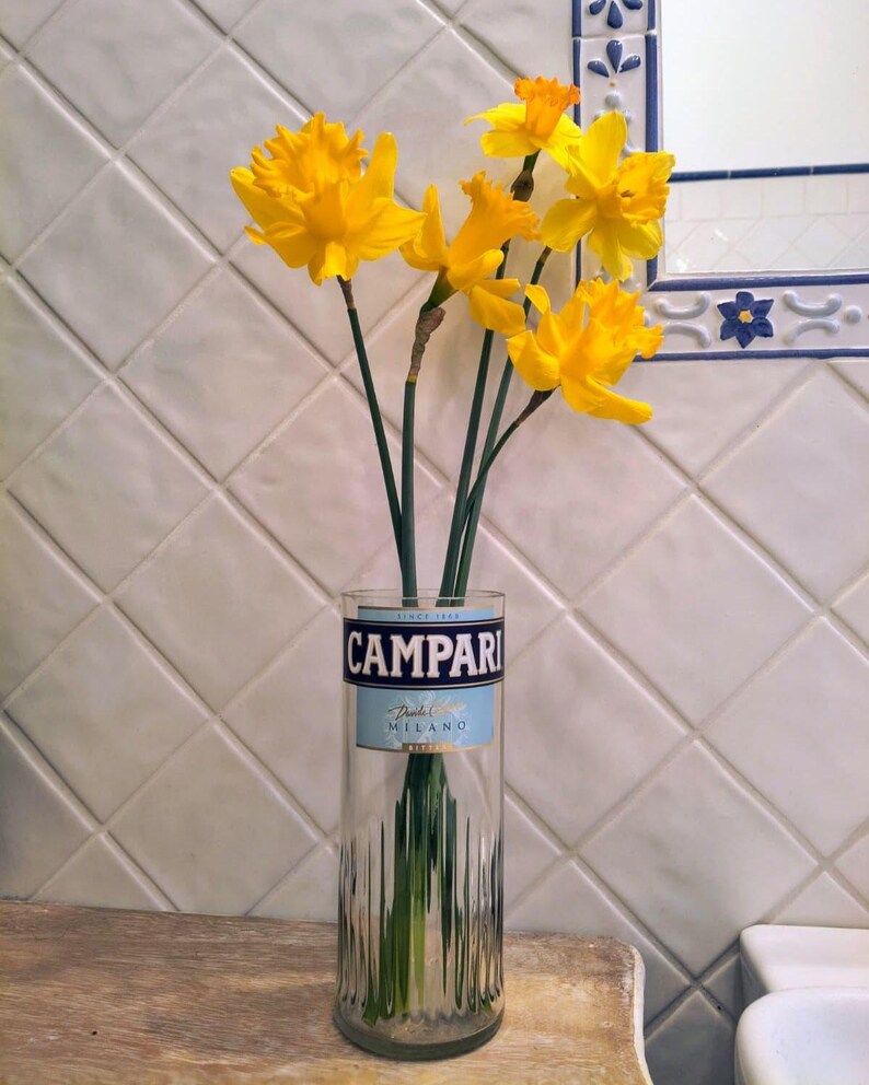 May include: A clear glass vase with a Campari label holds several bright yellow daffodils. The vase is on a wooden surface, and the background features white tiles and a mirror.