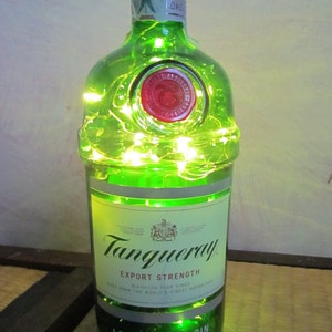 May include: A green glass Tanqueray gin bottle with a silver cap, red wax seal, and a white label. The bottle is illuminated with small, warm white fairy lights. The label reads "Tanqueray Export Strength London Dry Gin."