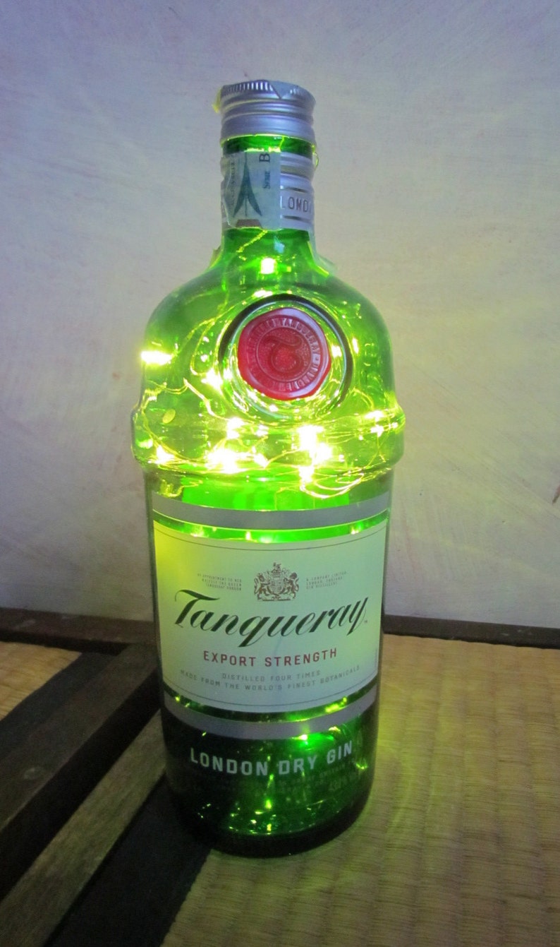 May include: A green glass Tanqueray gin bottle with a silver cap, red wax seal, and a white label. The bottle is illuminated with small, warm white fairy lights. The label reads "Tanqueray Export Strength London Dry Gin."