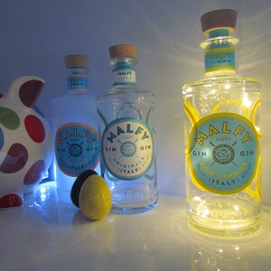 May include: Three bottles of Malfy Gin with blue and yellow labels, and a piggy bank with colorful polka dots. The gin bottles have cork stoppers and are illuminated with lights. One bottle reads "Con Limone".
