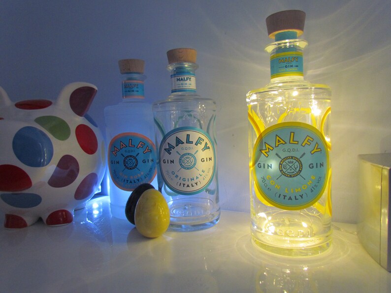 May include: Three bottles of Malfy Gin with blue and yellow labels, and a piggy bank with colorful polka dots. The gin bottles have cork stoppers and are illuminated with lights. One bottle reads "Con Limone".