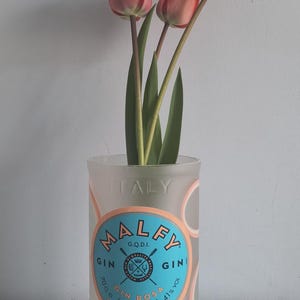May include: A clear glass bottle repurposed as a vase, holding three coral-colored tulips. The bottle features the text "MALFY GIN" and "ITALY" in a circular design with a blue and white label. The flowers have green stems and leaves.