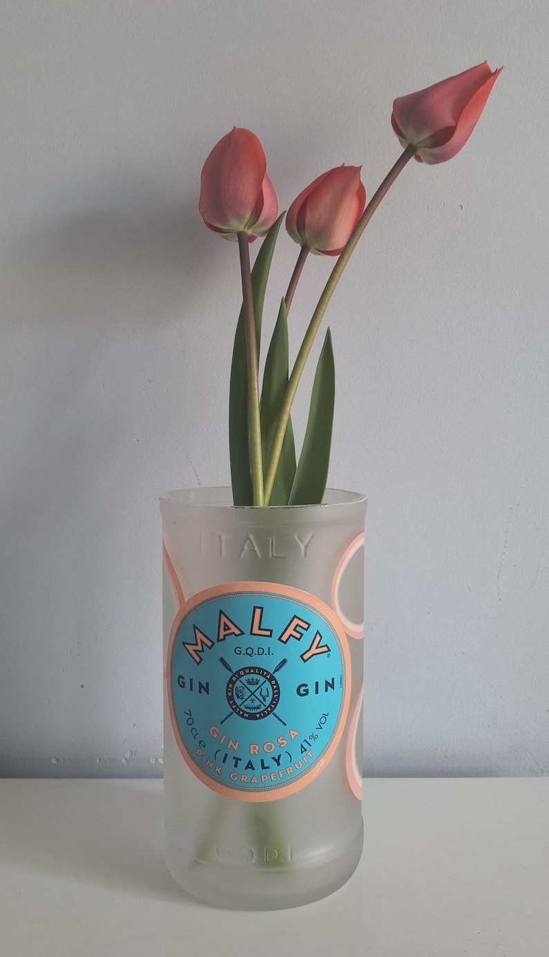 May include: A clear glass bottle repurposed as a vase, holding three coral-colored tulips. The bottle features the text "MALFY GIN" and "ITALY" in a circular design with a blue and white label. The flowers have green stems and leaves.