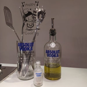 May include: A clear glass with the text "ABSOLUT VODKA" holds stainless steel kitchen utensils. A small bottle and a larger bottle of "ABSOLUT VODKA" are also present. The larger bottle contains a yellow liquid and has a metal spout.