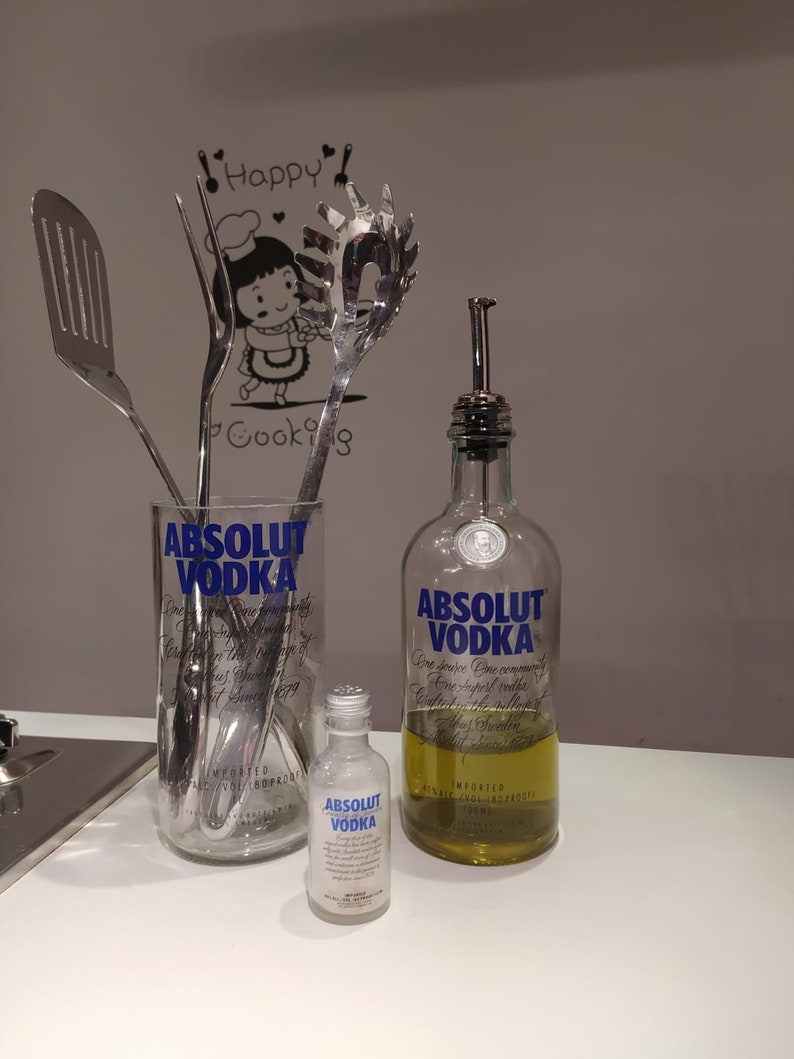 May include: A clear glass with the text "ABSOLUT VODKA" holds stainless steel kitchen utensils. A small bottle and a larger bottle of "ABSOLUT VODKA" are also present. The larger bottle contains a yellow liquid and has a metal spout.