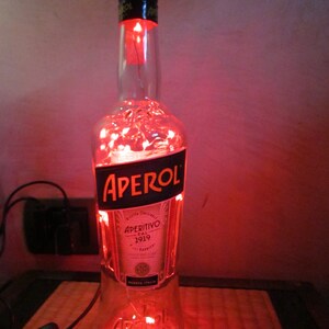 May include: A clear glass Aperol bottle with a black label and red string lights inside. The bottle is lit up with a warm red glow, creating a decorative lamp. The label reads "APEROL" and "APERITIVO DAL 1919".