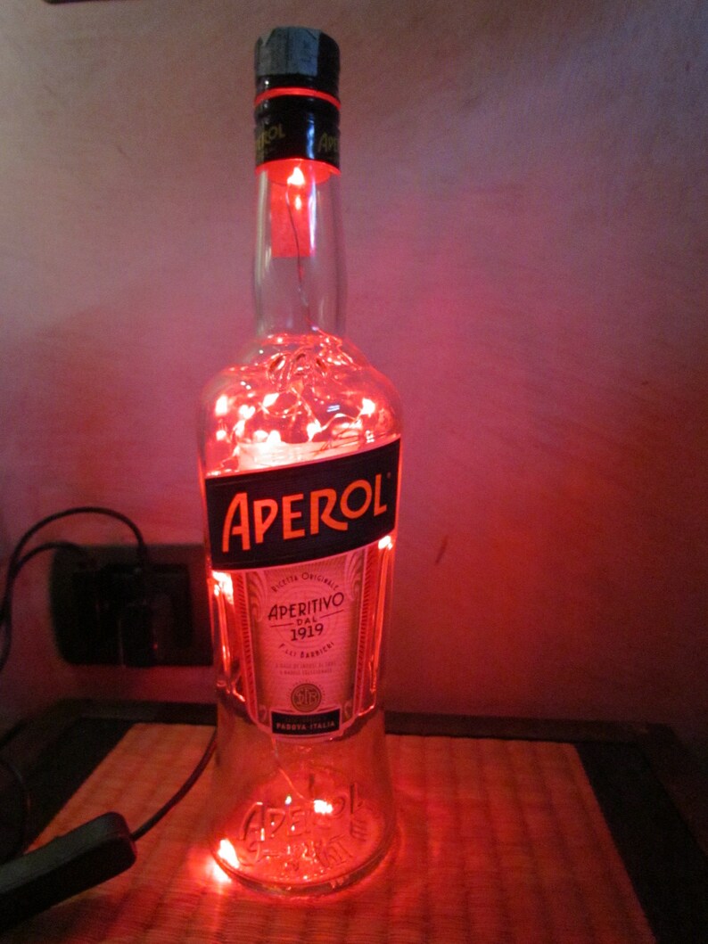 May include: A clear glass Aperol bottle with a black label and red string lights inside. The bottle is lit up with a warm red glow, creating a decorative lamp. The label reads "APEROL" and "APERITIVO DAL 1919".