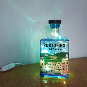 May include: A repurposed Portofino Dry Gin bottle illuminated with fairy lights. The bottle features a label with a colorful illustration of a coastal town. The bottle has a black cap and a white on/off switch.