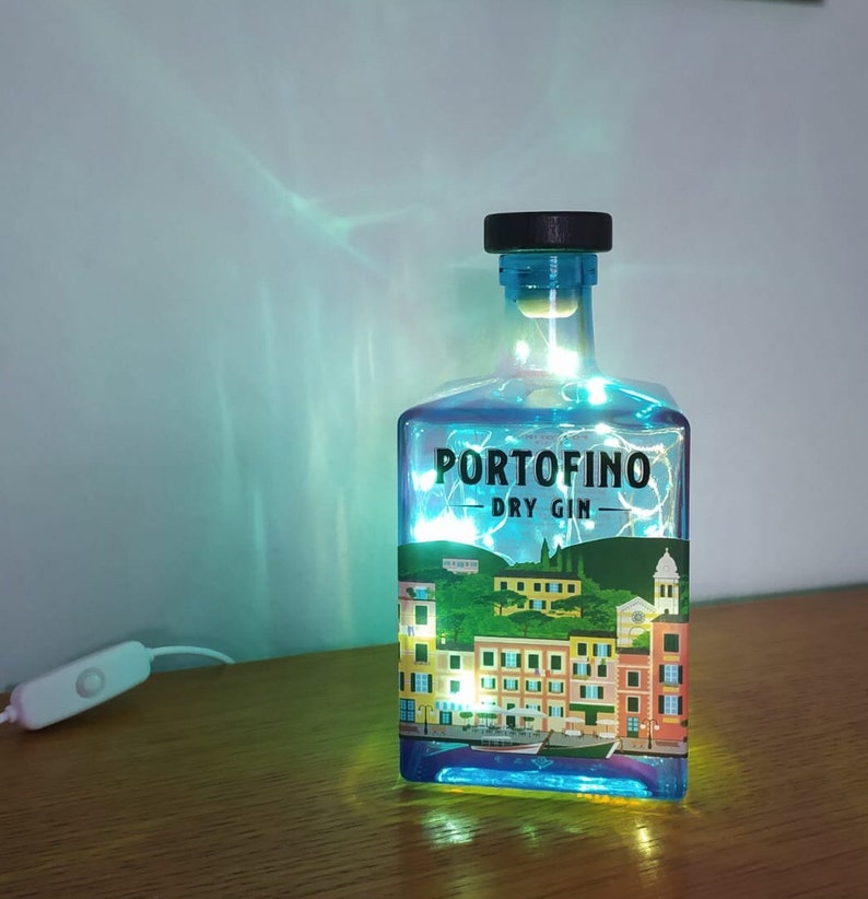 May include: A repurposed Portofino Dry Gin bottle illuminated with fairy lights. The bottle features a label with a colorful illustration of a coastal town. The bottle has a black cap and a white on/off switch.