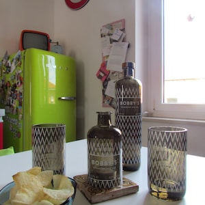 Puede incluir: Naturaleza muerta con una botella de Bobby's Schiedam Dry Gin, dos vasos a juego y una botella pequeña. Un cuenco de patatas fritas está delante. Las botellas y los vasos tienen un diseño geométrico. Un frigorífico verde retro está en el fondo.
