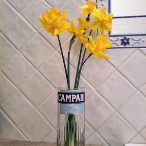 May include: A clear glass vase repurposed from a Campari bottle, holding a bouquet of bright yellow daffodils. The vase has a blue and white label that reads "CAMPARI" and "Da Milano". The flowers are in front of a white tiled wall.