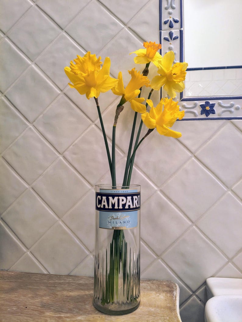 May include: A clear glass vase repurposed from a Campari bottle, holding a bouquet of bright yellow daffodils. The vase has a blue and white label that reads "CAMPARI" and "Da Milano". The flowers are in front of a white tiled wall.
