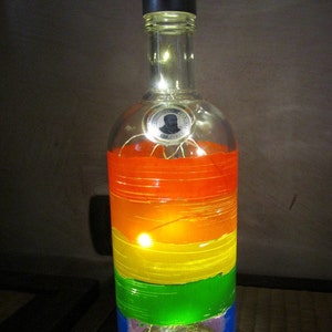 May include: A repurposed Absolut vodka bottle decorated with horizontal bands of rainbow colors. The bottle is illuminated with small fairy lights, creating a warm glow. The bottle has a black cap and a label with the text "Absolut Original Vodka".