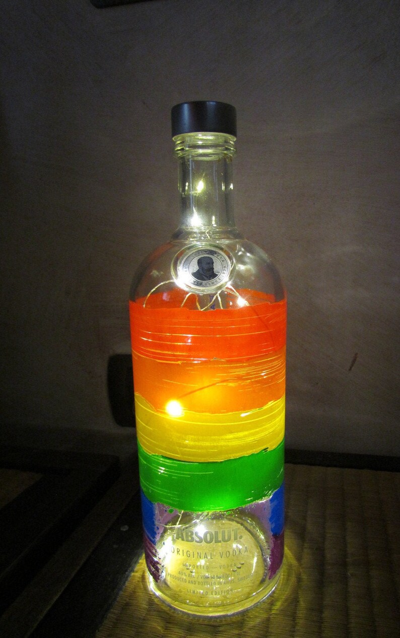 May include: A repurposed Absolut vodka bottle decorated with horizontal bands of rainbow colors. The bottle is illuminated with small fairy lights, creating a warm glow. The bottle has a black cap and a label with the text "Absolut Original Vodka".